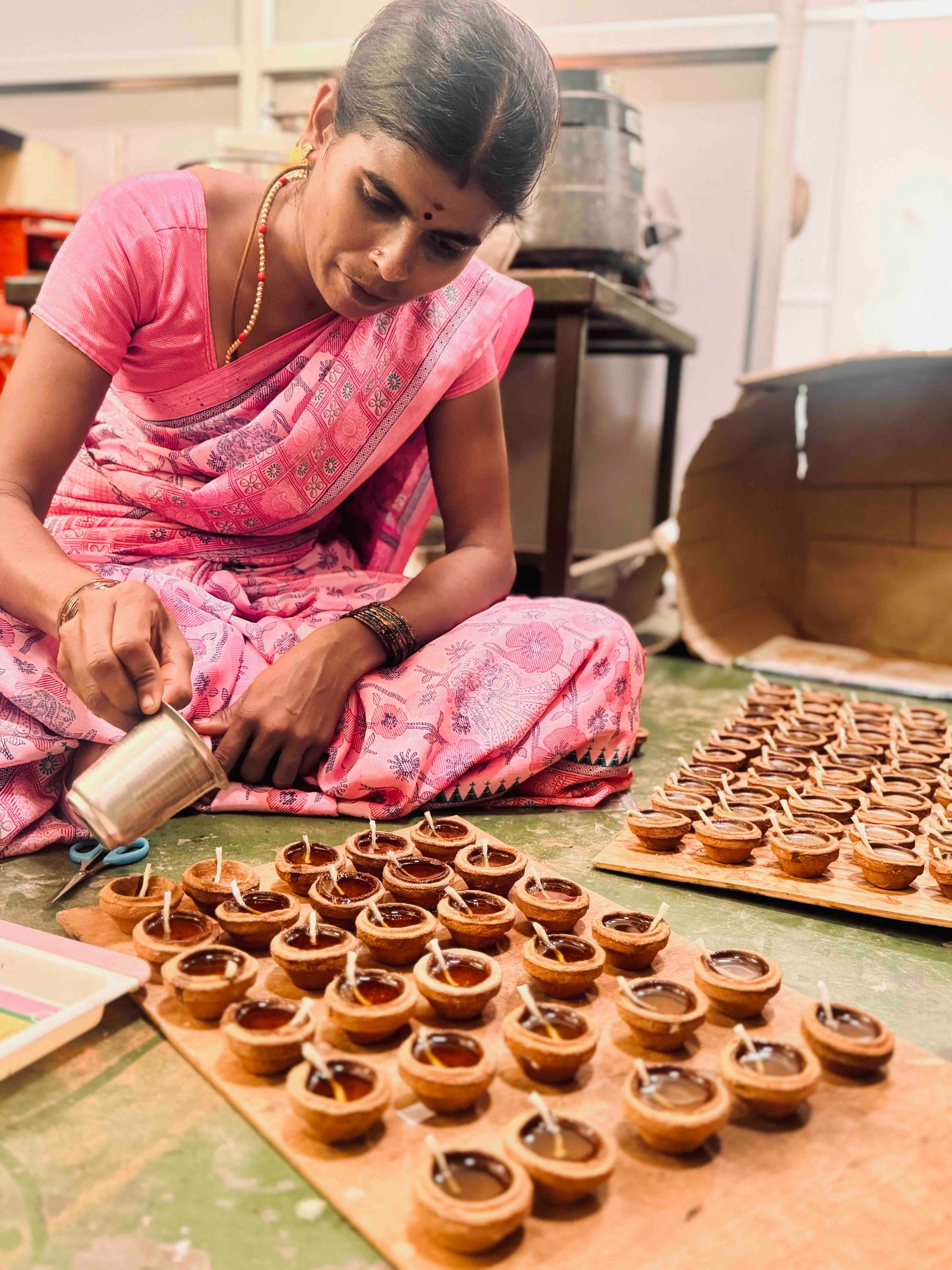 A woman in a pink sari filling rows of clay ghee lamps by hand on the PremaNature workshop floor