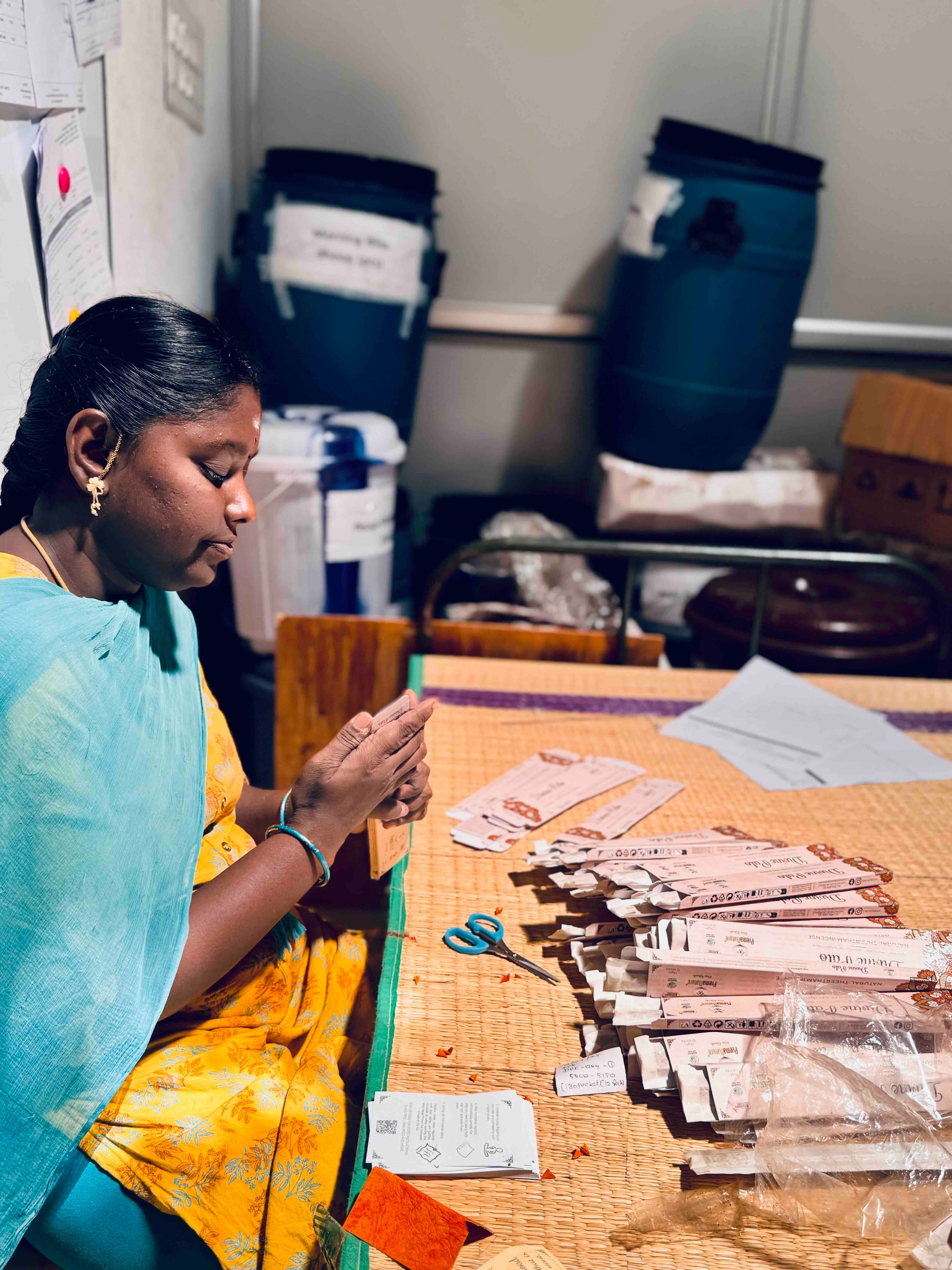 A woman in a yellow and turquoise sari labelling packs of PremaNature incense sticks by hand